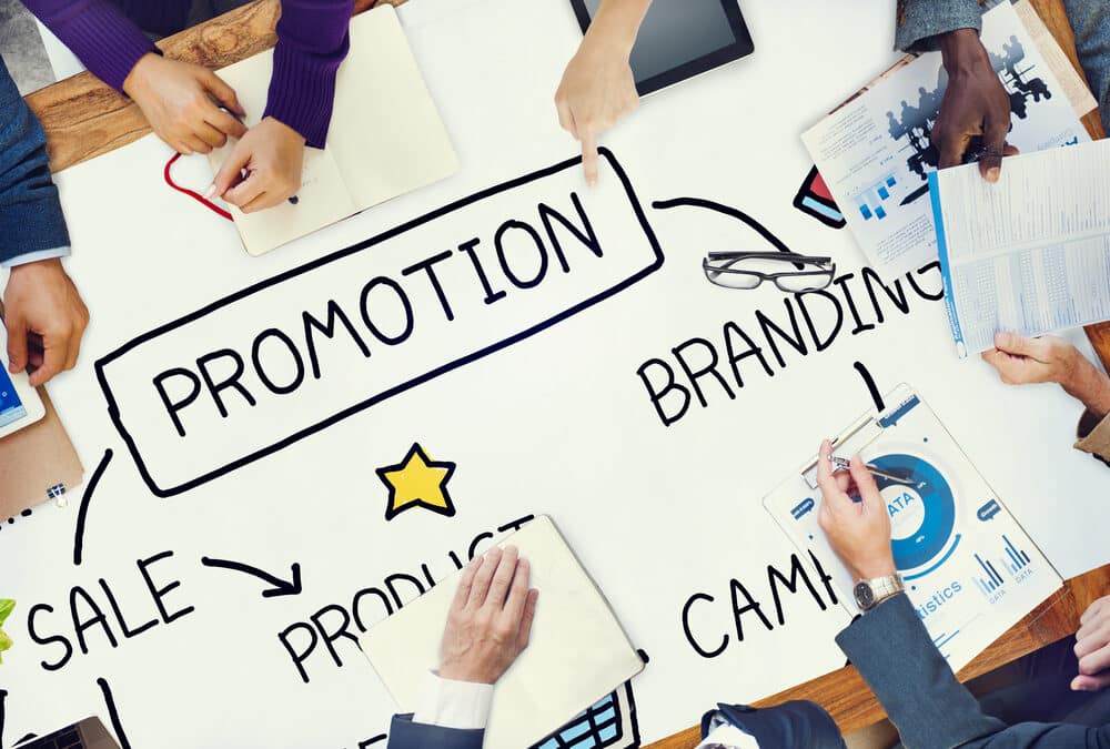 People sit around a table with a large paper showing a flowchart about promotion, including sale, branding, campaign, and product, with hands holding papers and devices.